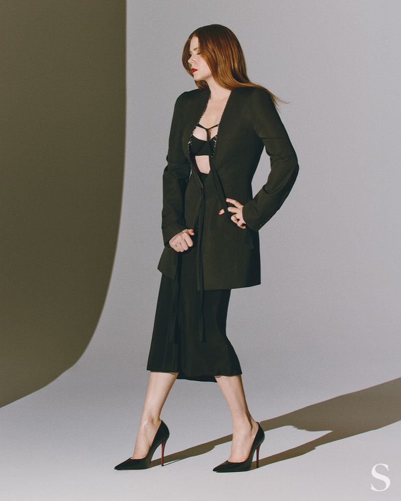 A redhead woman strikes a fashion pose in a blazer and heels in a studio setting with soft, directional lighting and a neutral color palette.