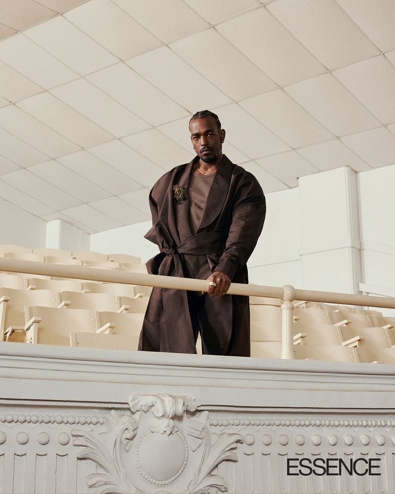 Man in a dark brown belted robe coat stands behind a theatre balcony railing with rows of empty auditorium seats behind him.