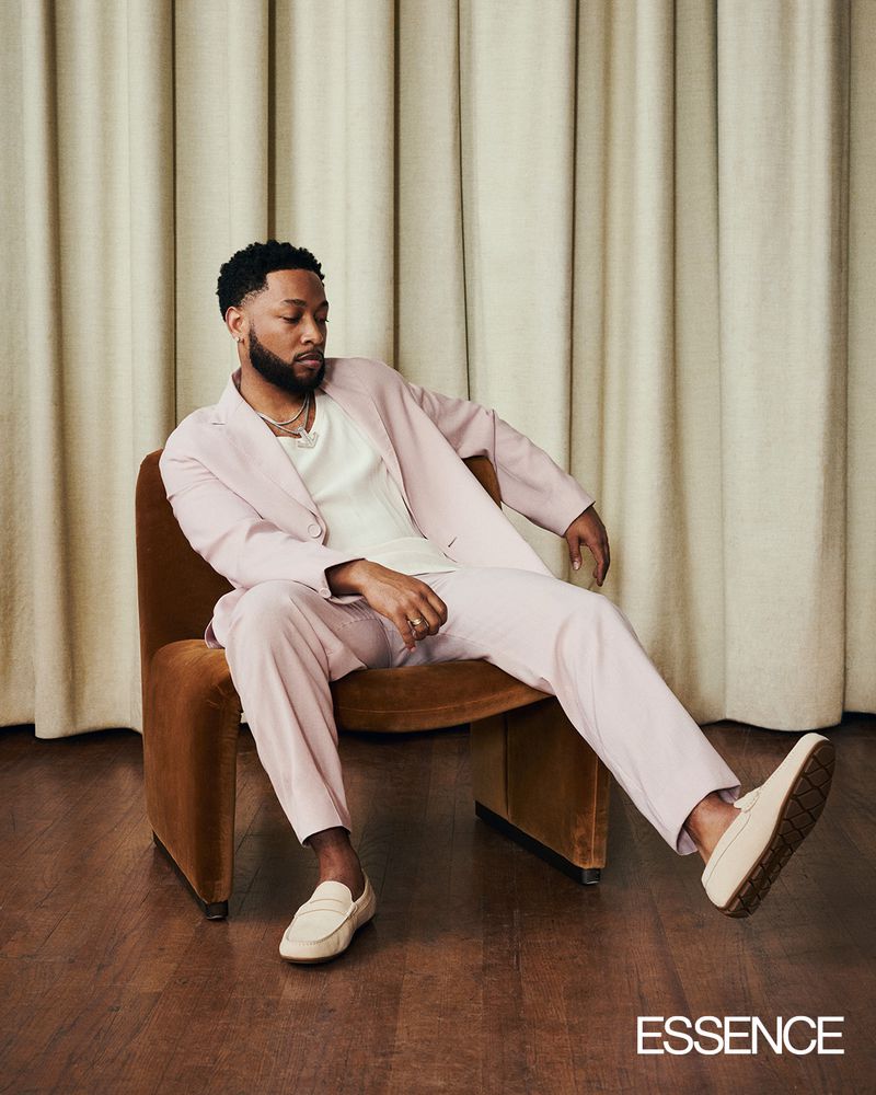 Black man in a light pink suit and cream loafers seated in a brown armchair against beige curtains.