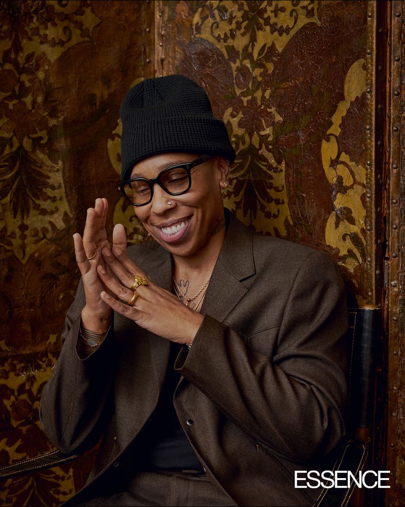 Lena Waithe smiling in a black beanie, eyeglasses, and brown blazer against a patterned backdrop.