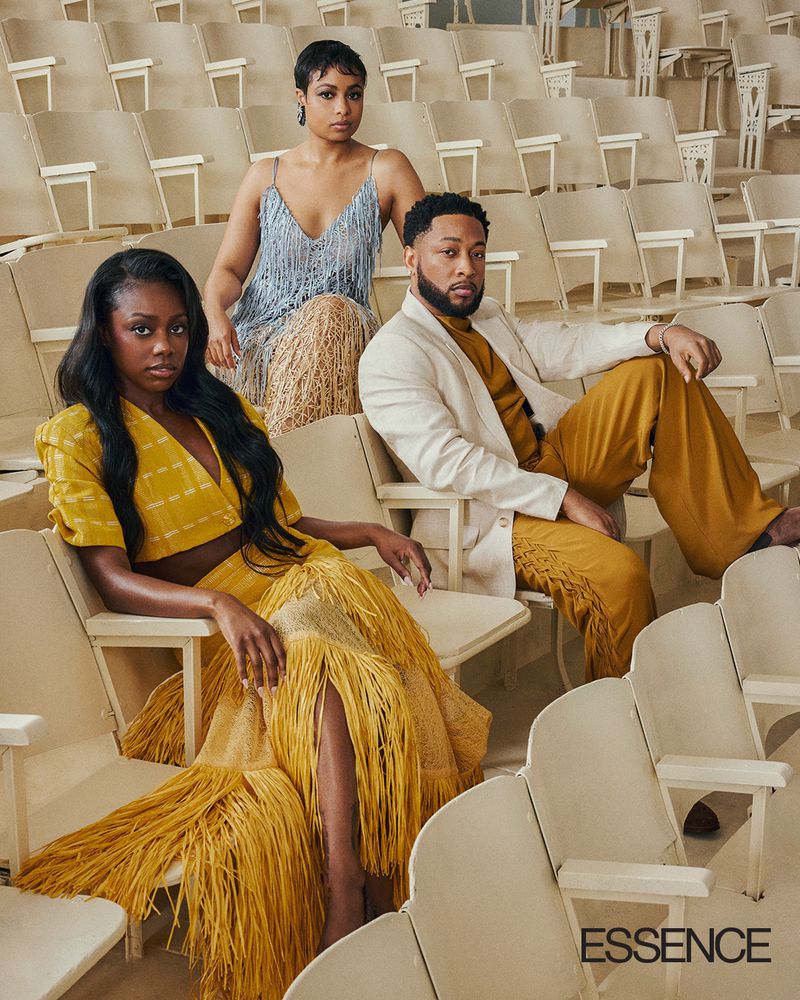 Group of three people in coordinated gold, yellow, and blue fringe outfits, seated in a theater setting in front of rows of empty seats.