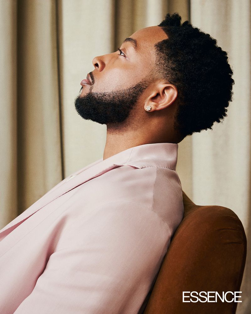 Profile portrait of a Black man wearing a pink blazer and diamond earring against draped curtains for Essence Magazine