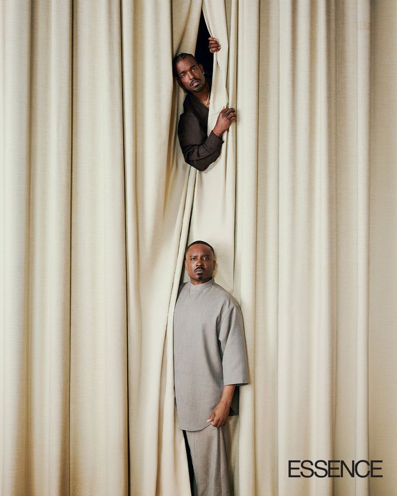 Two Black men looking out from between heavy vertical cream drapes in an editorial portrait for Essence Magazine.