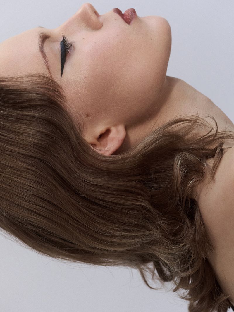 Side profile beauty portrait of a woman with dramatic black winged eyeliner and wavy light brown hair against a pale neutral background.