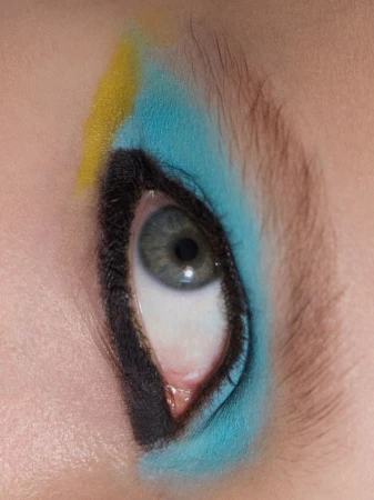 Extreme close-up of a blue-grey eye with bold black eyeliner, vibrant sky blue eyeshadow, and a streak of yellow makeup.