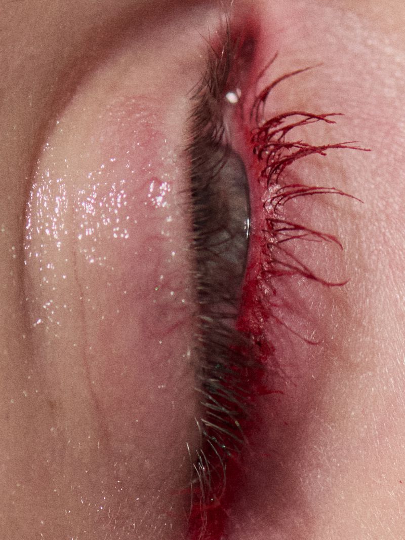 Extreme close-up of an eye featuring vibrant red mascara on individual eyelashes with glossy eyelid skin texture.