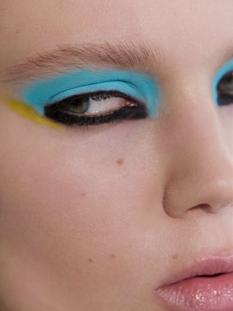Close-up of a woman with bold graphic eye makeup featuring vibrant blue eyeshadow, thick black liner, and a yellow accent wing.