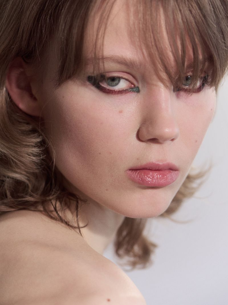 Close-up portrait of a woman with bangs and bold graphic eyeliner looking toward the camera against a neutral studio backdrop.