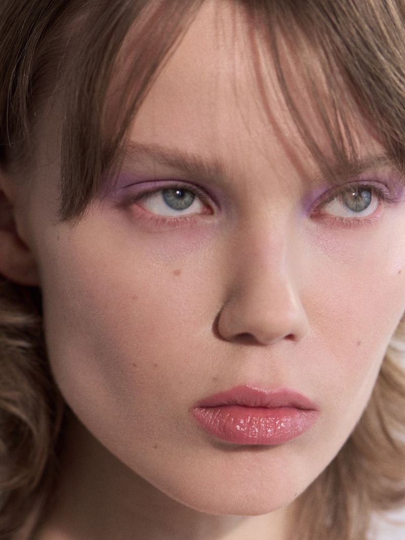 Close-up beauty portrait of a woman with lilac eyeshadow, glossy pink lips, and wispy bangs looking upward.