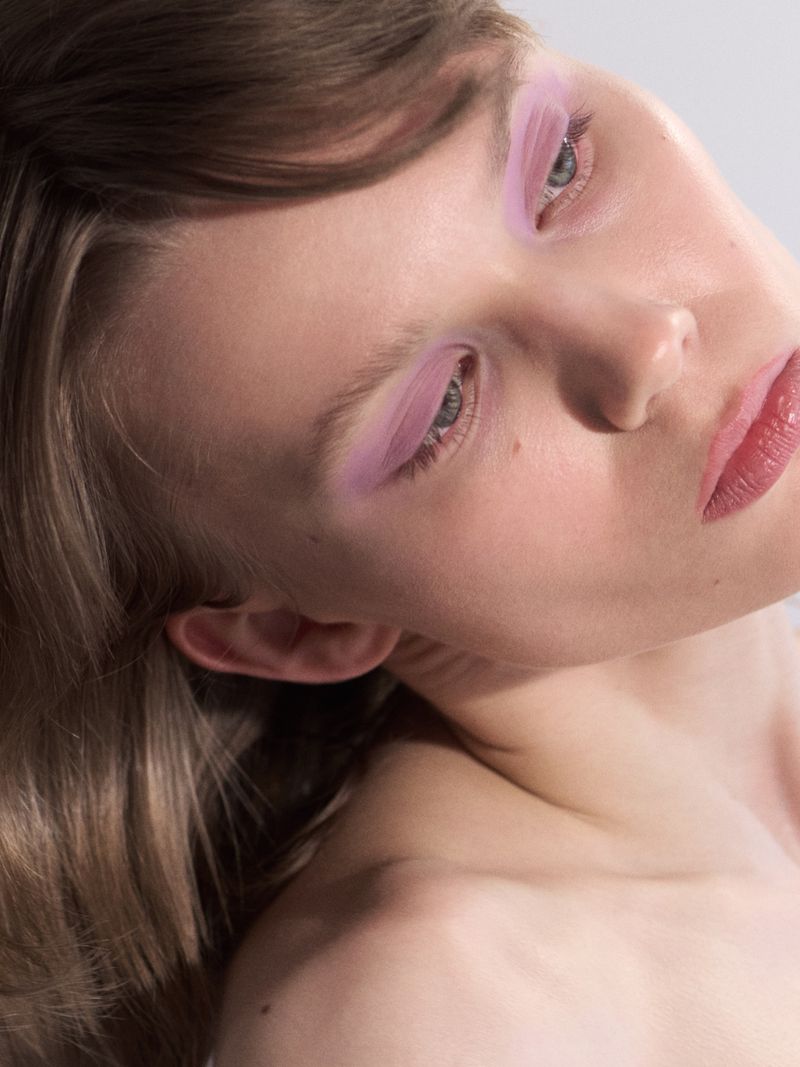 Close-up beauty portrait of a woman tilting her head with pastel pink eye makeup and bare shoulders against a neutral backdrop.