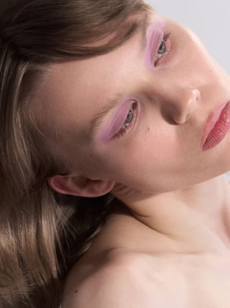 Close-up beauty portrait of a woman tilting her head with pastel pink eye makeup and bare shoulders against a neutral backdrop.