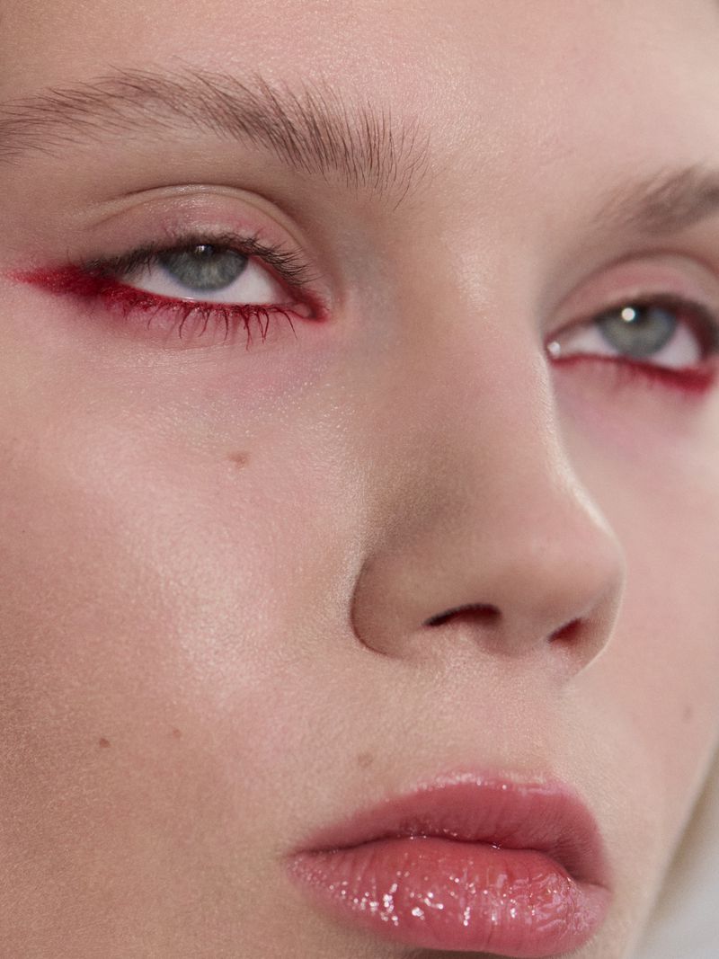 Close-up beauty portrait showing red winged eyeliner, natural brows, and glossy pink lips on light skin.