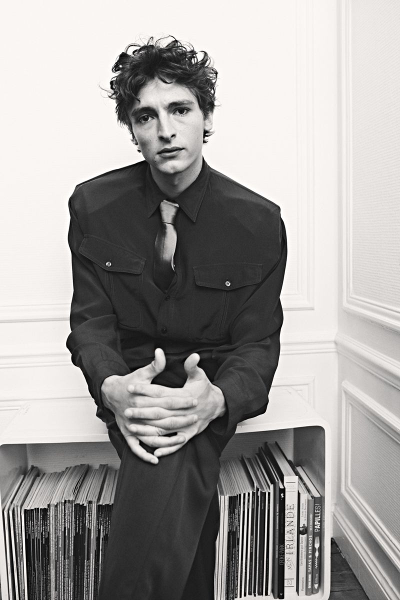 Black and white portrait of Vassili Schneider in a dark button-up shirt and tie, sitting on a low bookshelf.