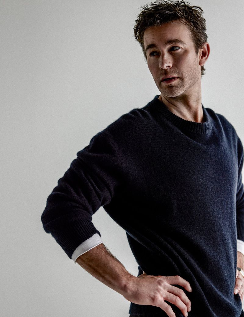 Actor Chace Crawford posing in a dark navy blue crewneck knit sweater against a light grey seamless studio background.