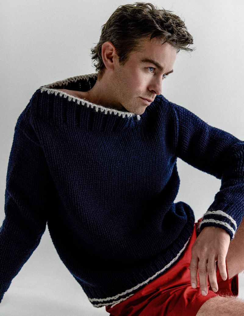 Chace Crawford wearing a navy blue knit sweater with white trim and red shorts against a plain white studio background.