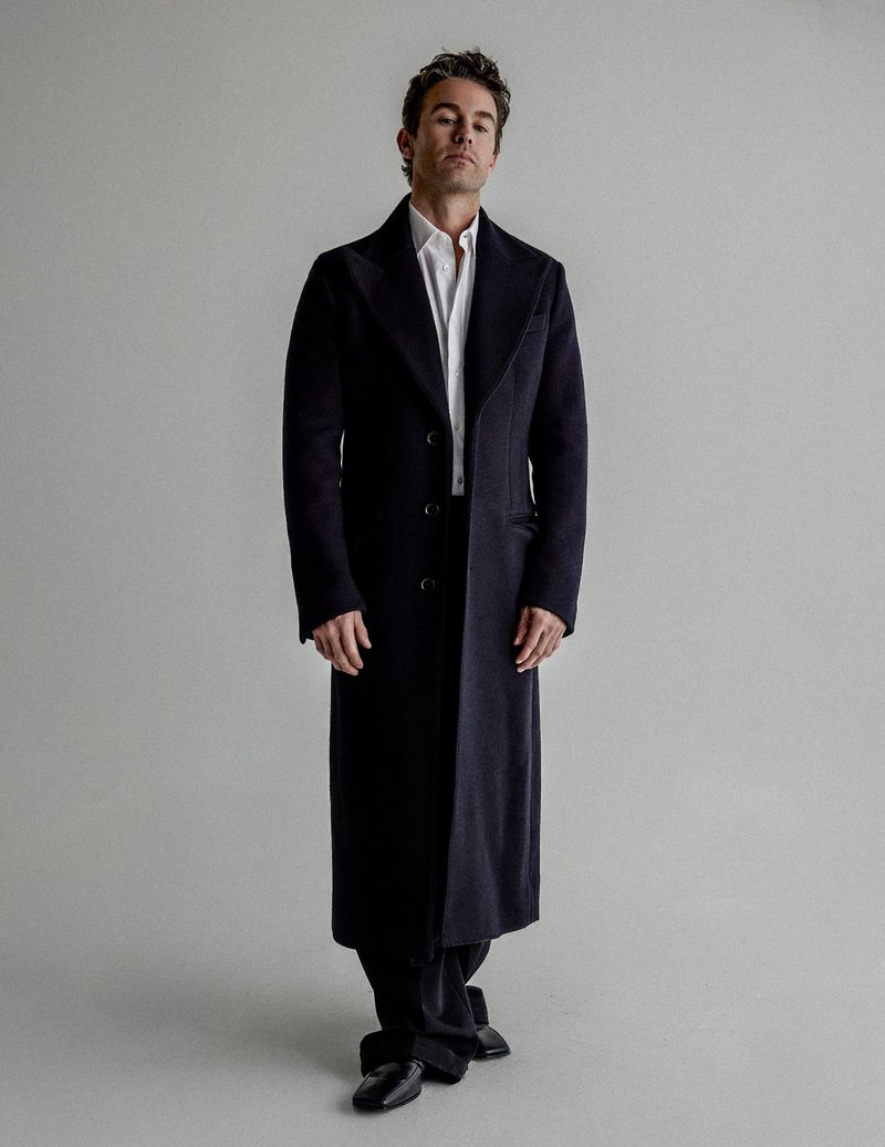 Chace Crawford posing in a long dark tailored overcoat, white dress shirt, and black trousers against a neutral grey studio backdrop.