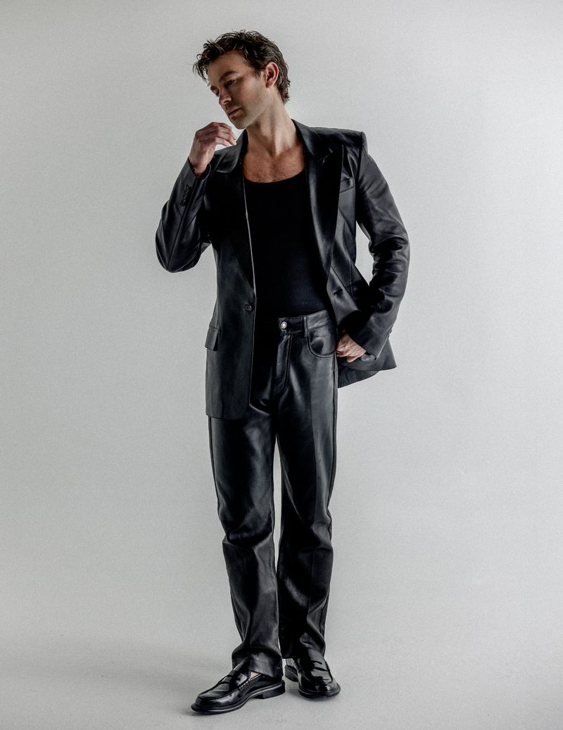 Full-length portrait of Chace Crawford wearing a black leather blazer and matching pants against a plain studio background.