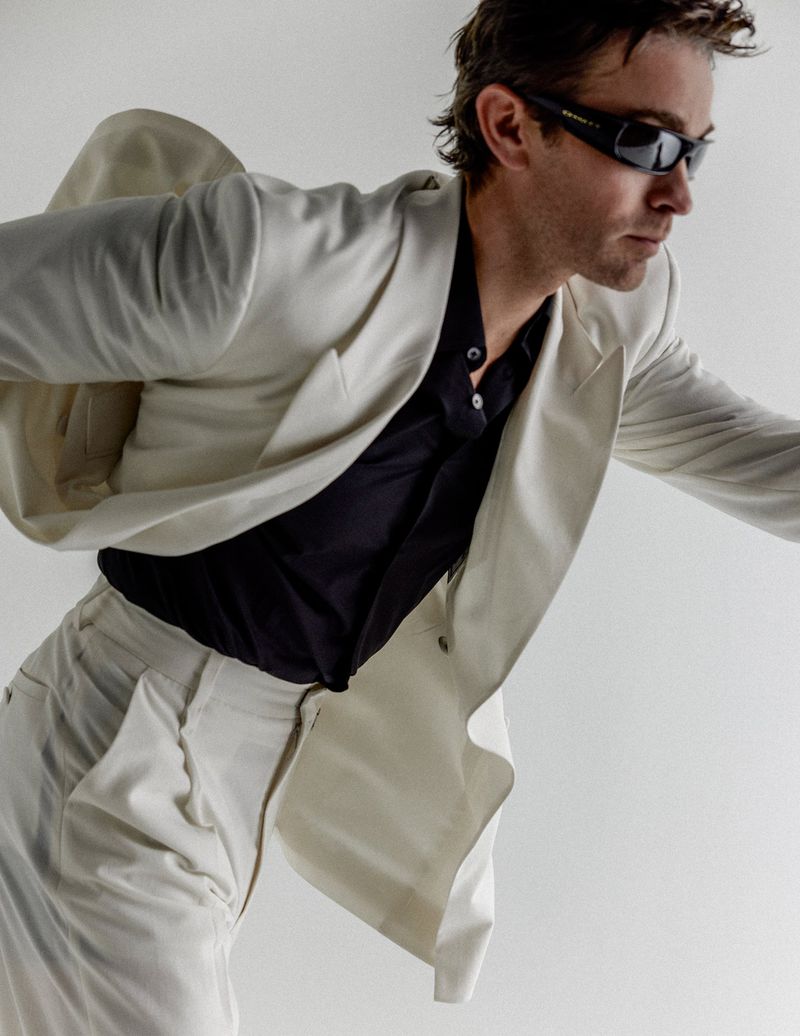 Chace Crawford wearing dark sunglasses and an off-white blazer over a black shirt against a white seamless background.
