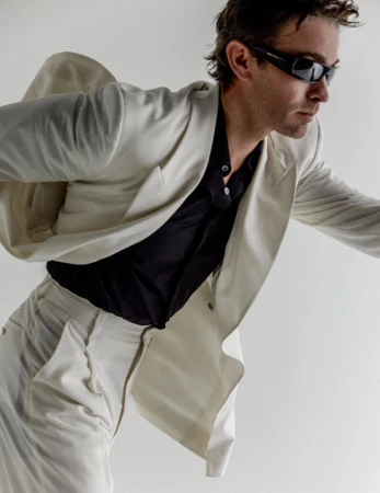 Chace Crawford wearing dark sunglasses and an off-white blazer over a black shirt against a white seamless background.