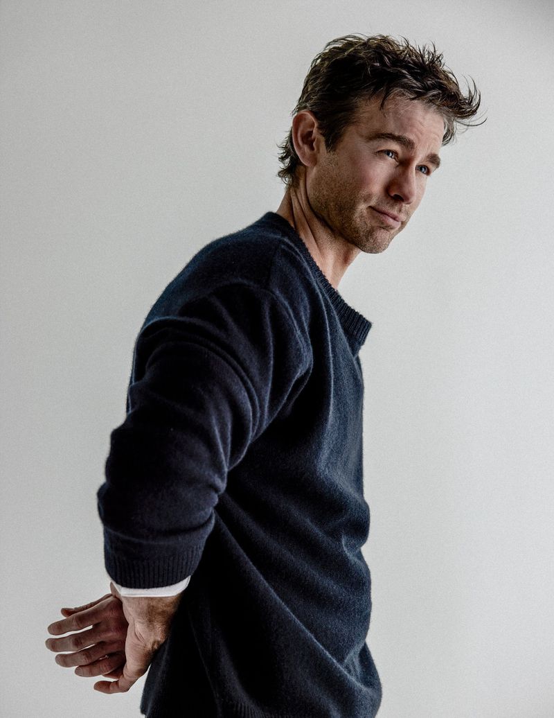 Chace Crawford standing in profile wearing a dark navy sweater with hands behind his back against a white studio background.