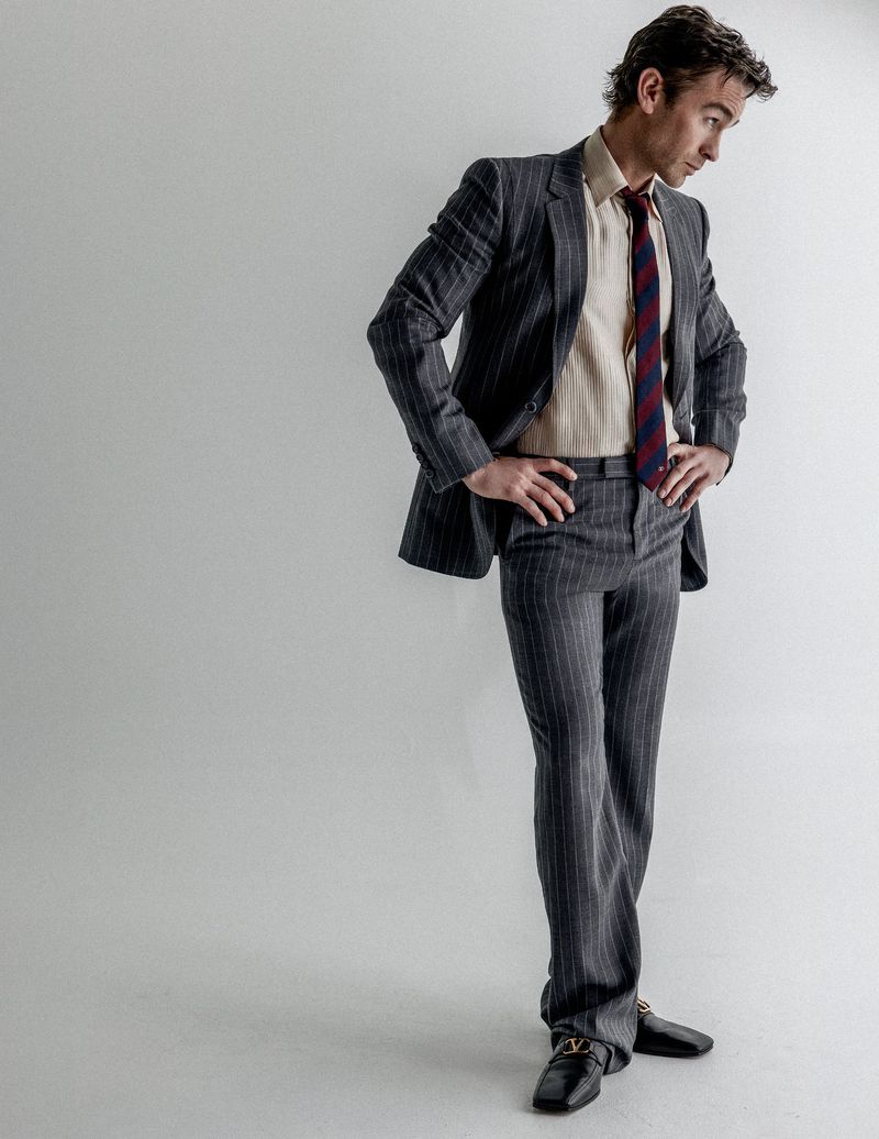 Chace Crawford wearing a tailored grey pinstripe suit and striped tie against a white studio backdrop.