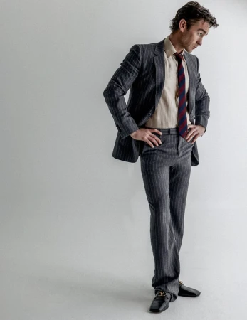 Chace Crawford wearing a tailored grey pinstripe suit and striped tie against a white studio backdrop.