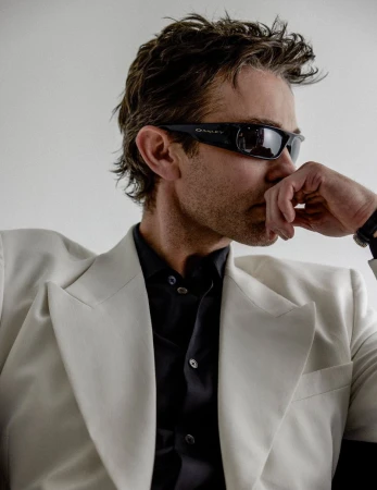 Chace Crawford wearing black wrap sunglasses and an off-white blazer with peak lapels over a dark button-up shirt in profile pose.