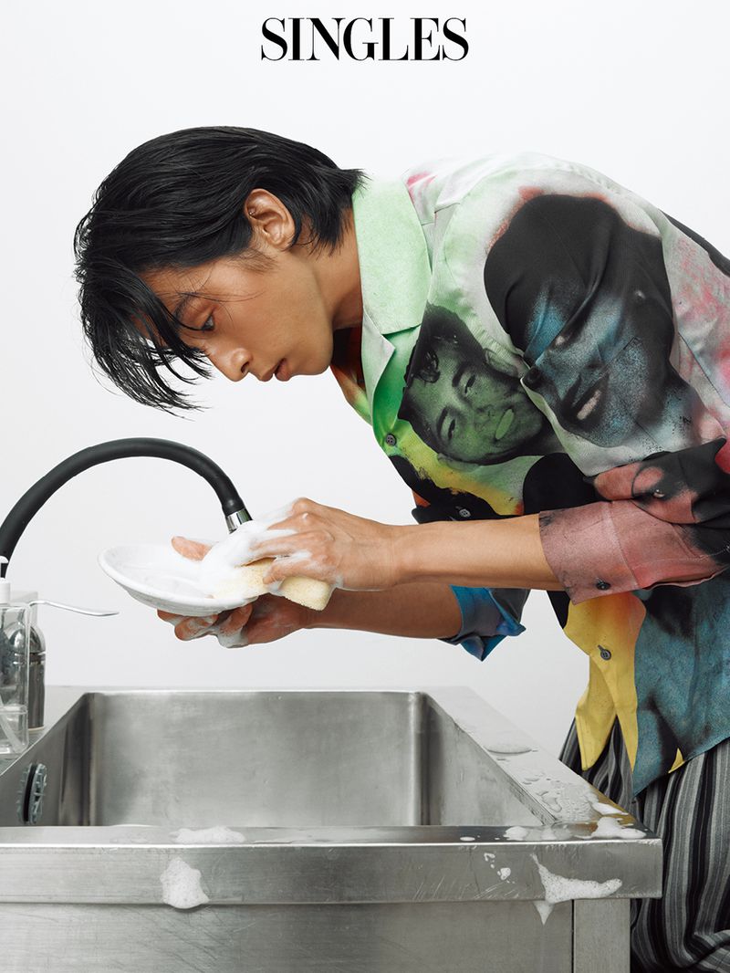 Model Yugo Takano washing dishes with soapy foam over a stainless steel sink in a graphic printed shirt against a white backdrop.