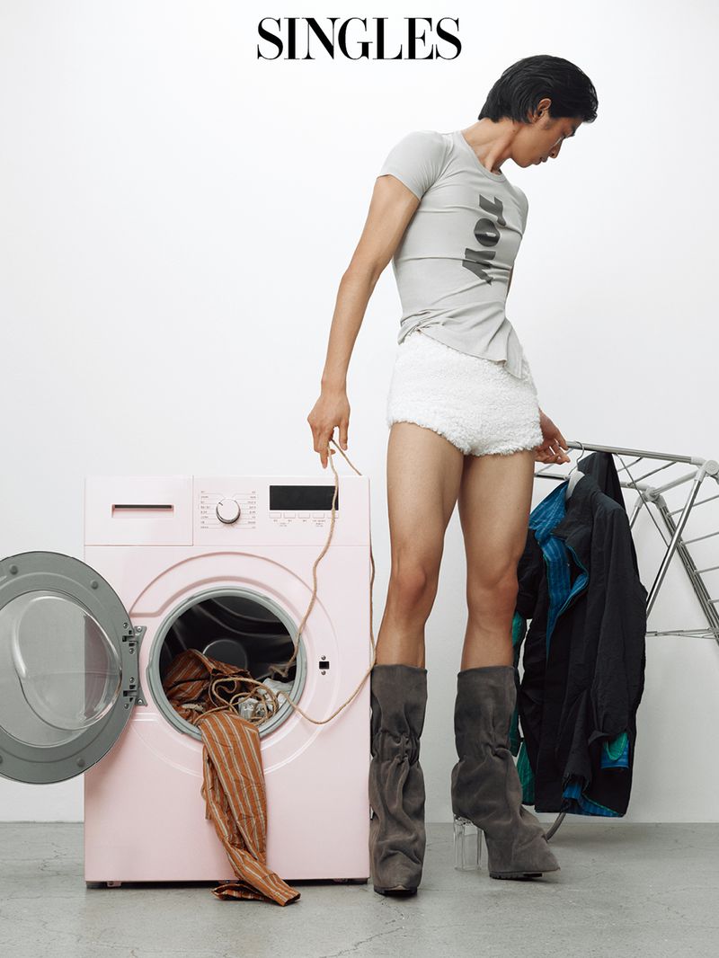 Fashion editorial shot of a slender Asian male in a gray t-shirt, white shorts, and gray boots, standing next to a pink washing machine.