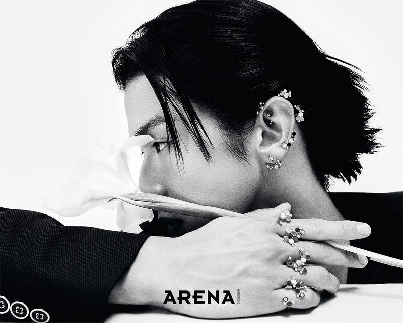 Monochrome studio portrait of an Asian adult with short dark hair, wearing rings and earrings, and holding a white flower.