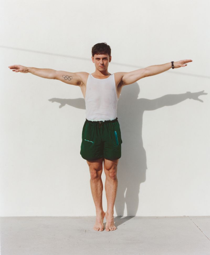 Tom Daley standing against a white wall with arms outstretched, wearing a white tank top and dark green shorts.