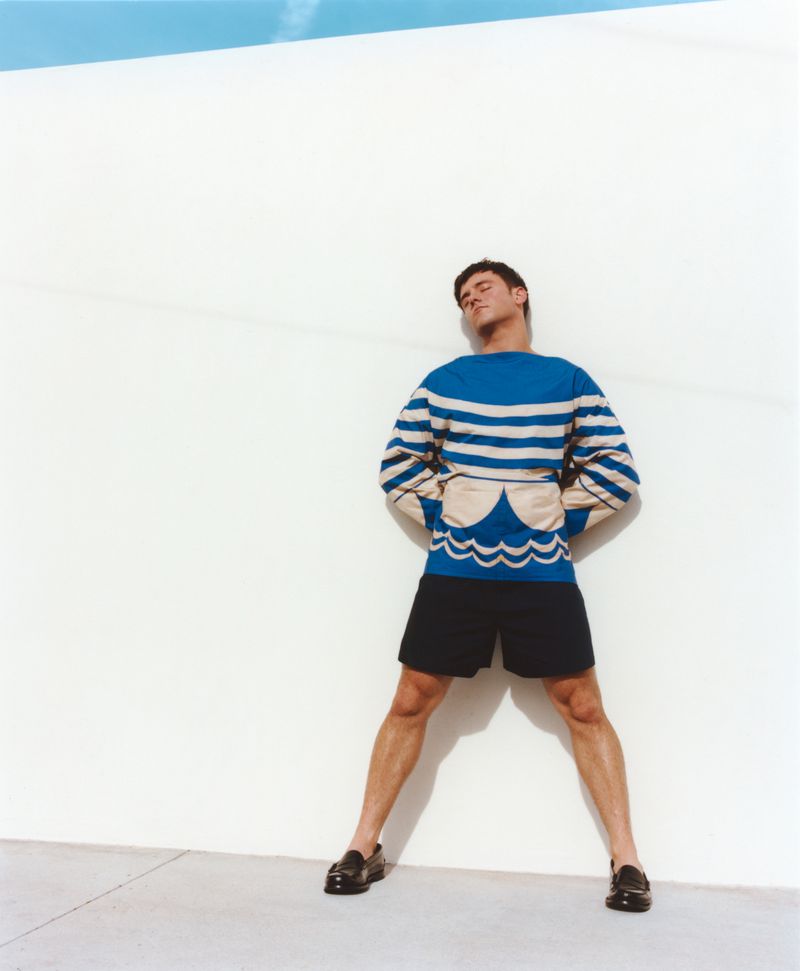 Athlete Tom Daley posing against a white wall wearing a blue and white patterned sweater and dark shorts