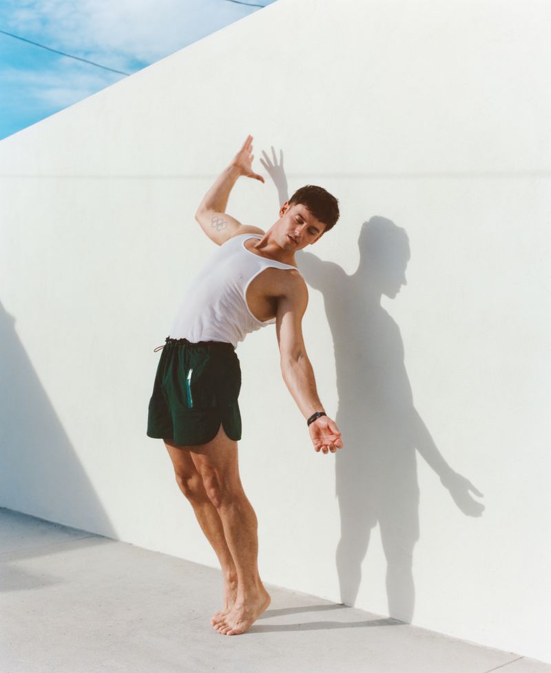 Tom Daley in a white tank top and dark green shorts posing against a white wall for Behind the Blinds.