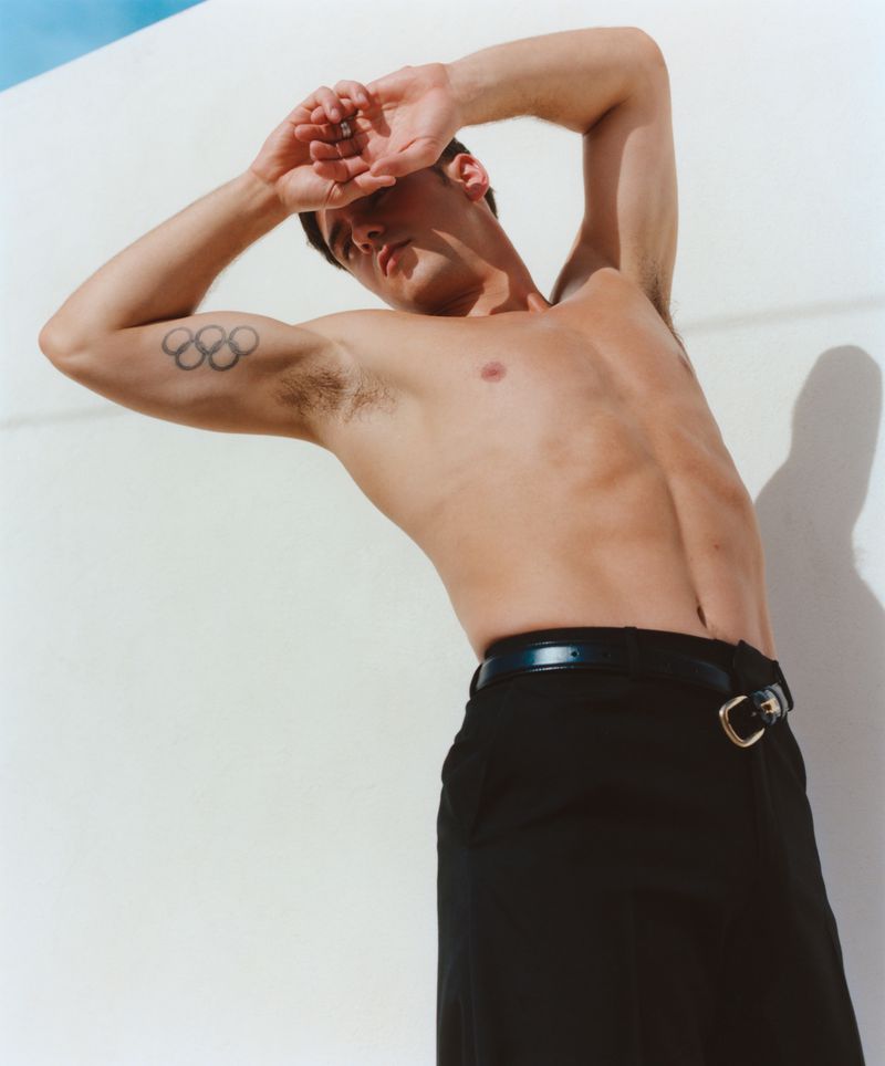 Portrait of Tom Daley shirtless wearing black trousers with arms raised behind his head against a white background.