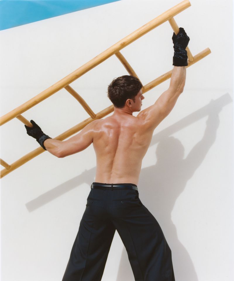 Male model in black pants and gloves posing with a wooden ladder against a white wall with a blue accent, showcasing fitness and fashion.