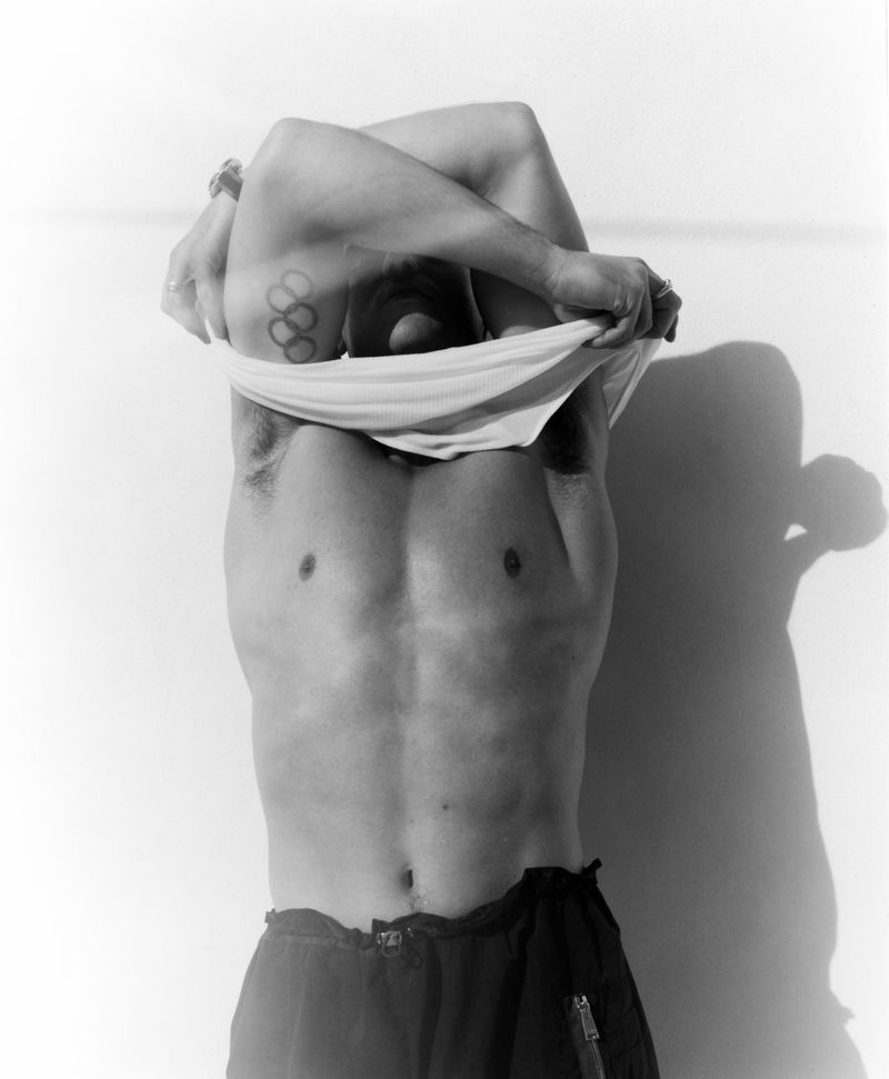 Tom Daley taking off a white t shirt in a black and white editorial portrait against a plain studio background.