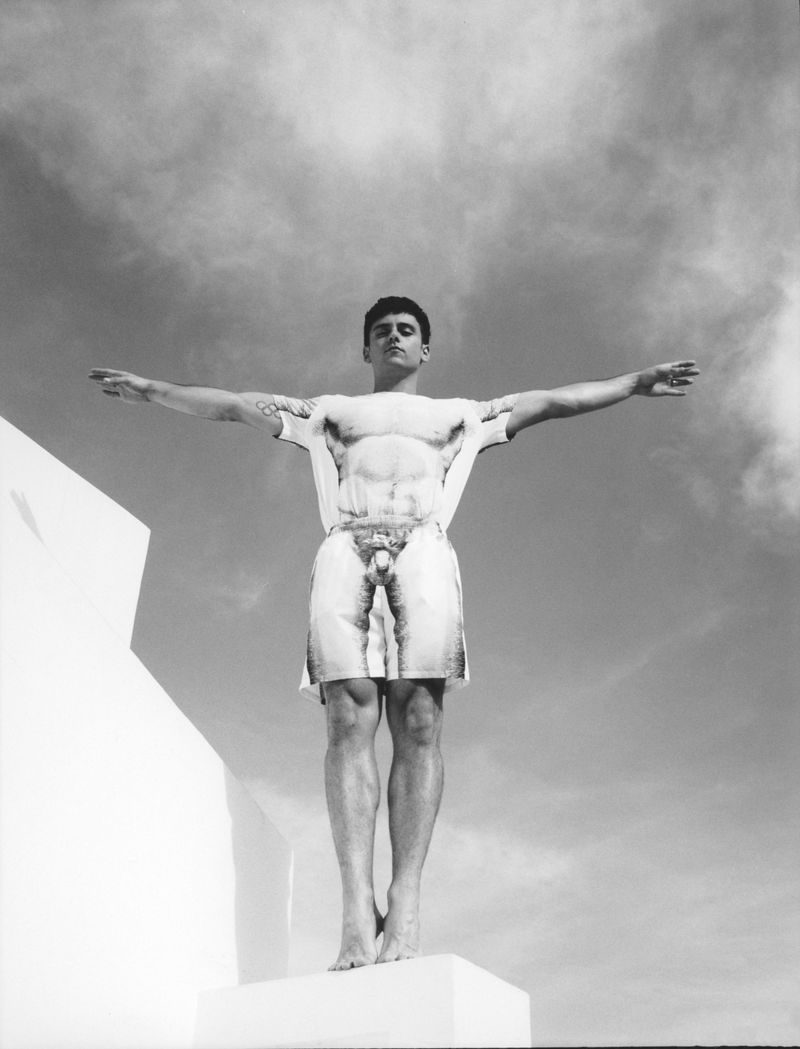Monochrome portrait of Tom Daley standing with arms outstretched on a white architectural pedestal against a cloudy sky.