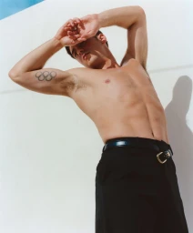 Portrait of Tom Daley shirtless wearing black trousers with arms raised behind his head against a white background.
