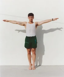Tom Daley standing against a white wall with arms outstretched, wearing a white tank top and dark green shorts.