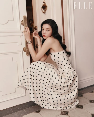 A woman with dark hair poses in a white strapless polka-dot dress with a cinched waist, in an indoor setting with ornate decor.