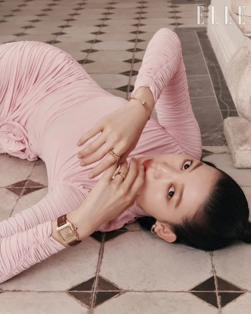 Asian female model in a long-sleeve pink dress lies on a patterned tile floor, accessorized with jewelry and a brown watch in a studio.