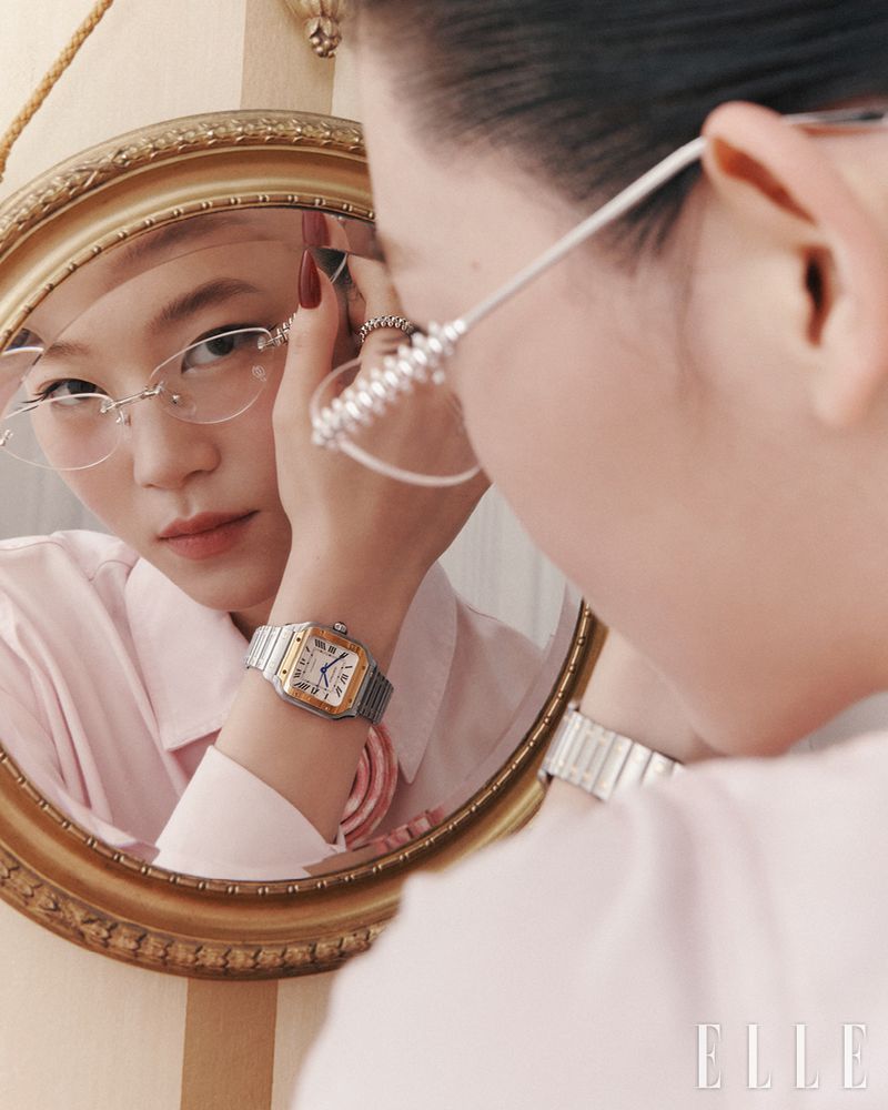 Asian woman with glasses looks in a round mirror with a gold frame, wearing a watch, a pink shirt, and ring on her manicured hand.