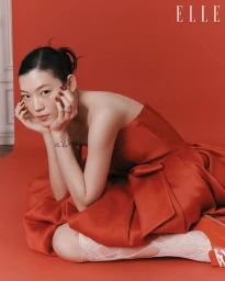 Model wearing a red dress and wrist watch seated against a red studio backdrop for Elle Korea editorial.