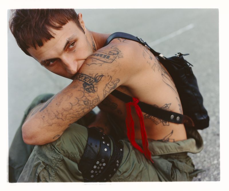 Anwar Hadid seated outdoors wearing green cargo trousers and a black strap bag, showcasing numerous tattoos.