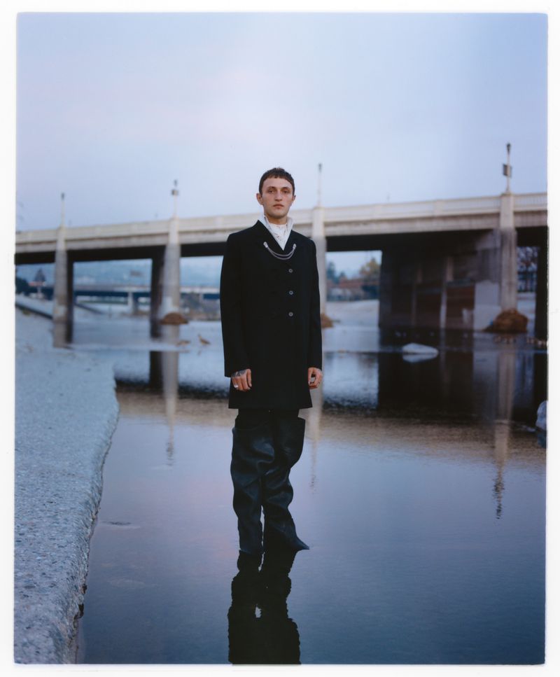 Model Anwar Hadid stands in shallow river water wearing a black coat and tall waders beneath a concrete bridge.