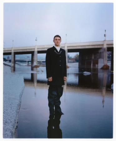 Model Anwar Hadid stands in shallow river water wearing a black coat and tall waders beneath a concrete bridge.