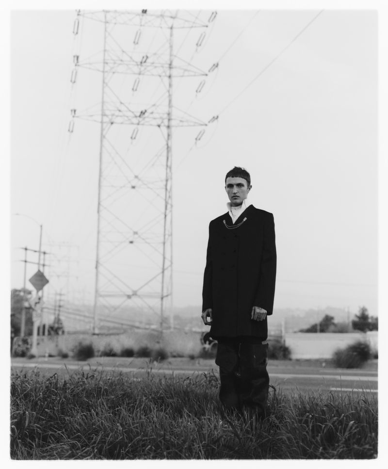 Black and white portrait of Anwar Hadid standing in tall grass wearing a dark coat in front of a large electrical transmission tower.