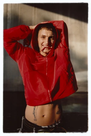 Anwar Hadid wearing a hooded red nylon windbreaker with hands behind head and visible torso tattoos outdoors.