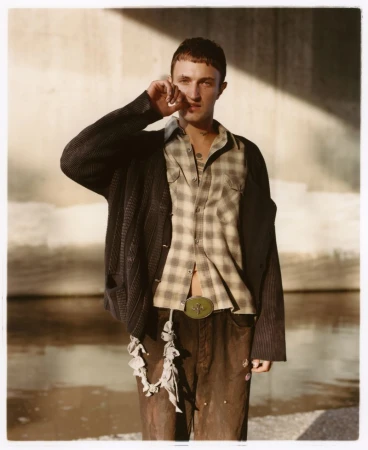 Anwar Hadid wearing an open plaid shirt and dark cardigan, standing in direct sunlight against a concrete wall and canal.