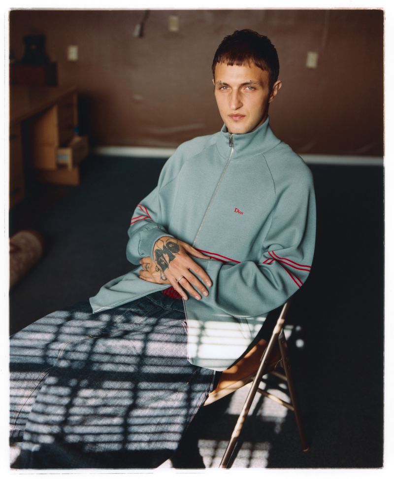 Anwar Hadid seated on a folding chair wearing a light blue zip-up jacket and dark trousers for Behind the Blinds.